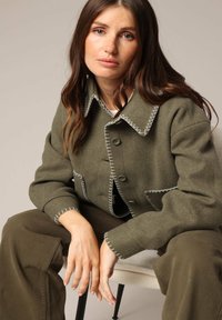 Veste en laine vert olive avec des coutures blanches contrastantes, grands revers et poches avant. Le modèle porte un pantalon assorti et est assis sur une chaise.