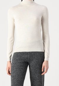 Kvinna som bär en ljusbeige polotröja och mörkgrå strukturerade byxor, visad från halsen till mitten av låret mot en enkel bakgrund.