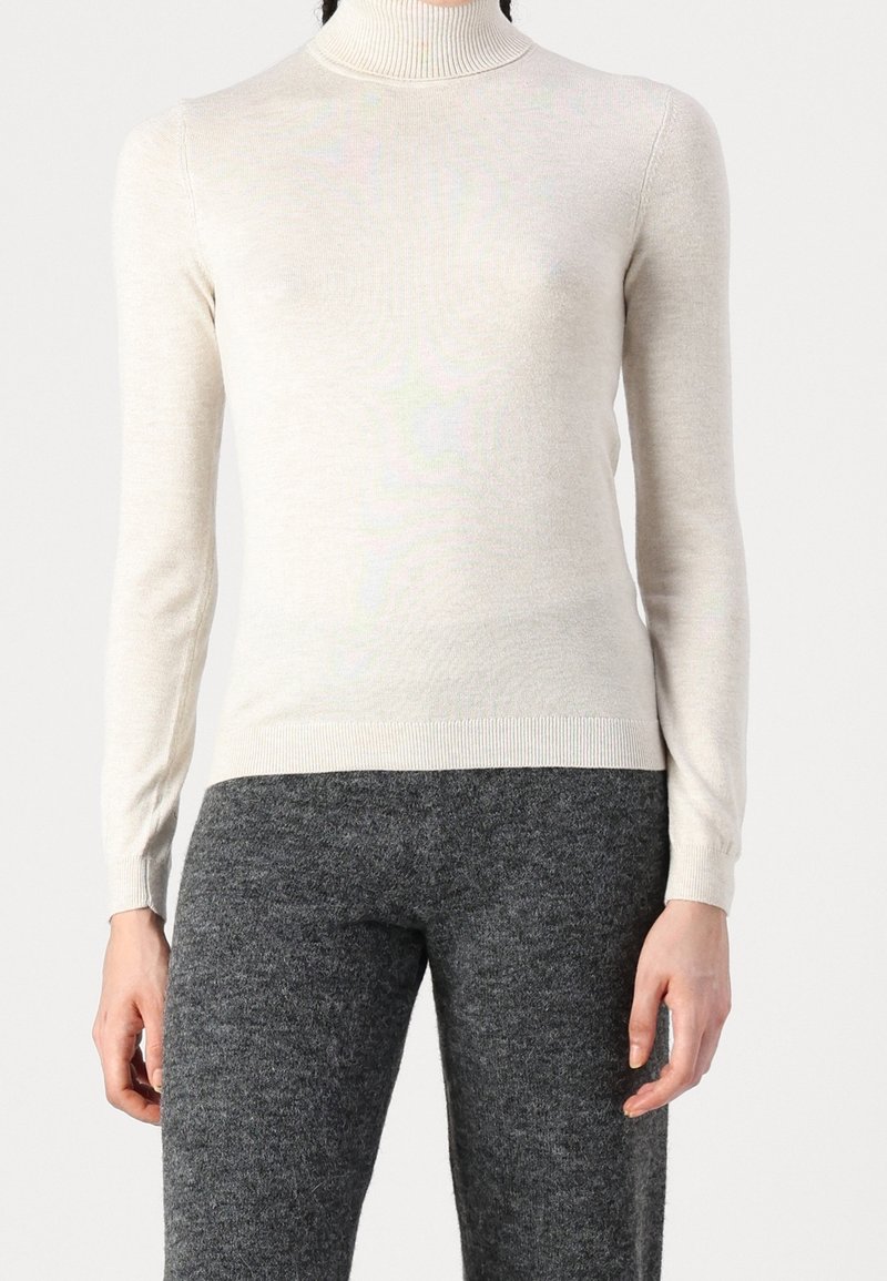 Kvinna som bär en ljusbeige polotröja och mörkgrå strukturerade byxor, visad från halsen till mitten av låret mot en enkel bakgrund.