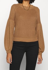 Stickad tröja i ljusbrun med ribbad textur, raglanärmar och smockade manschetter. I kombination med svarta byxor och ett guldhalsband.