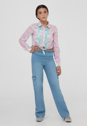 Camicia a maniche lunghe, con bottoni, in un pattern tie-dye rosa, blu e verde, annodata in vita, abbinata a jeans cargo azzurri chiari con tasche.