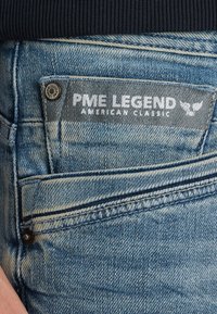 Ljusblå denimjeans med en grå tygetikett med "PME LEGEND AMERICAN CLASSIC" i vit skrift, detalj av framfickan synlig.