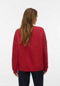 Rdeč pulover z ohlapnim krojem, dolgimi rokavi in rebrastimi manšetami; gladka tekstura in klasičen okrogel izrez; nošen s temnimi jeansa hlačami.