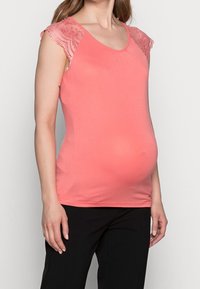 Korálový těhotenský top s krajkovými zavinovacími rukávy, vykrojeným výstřihem a elastickým materiálem. Jsou na sobě černé kalhoty, které zvýrazňují přiléhavou siluetu.