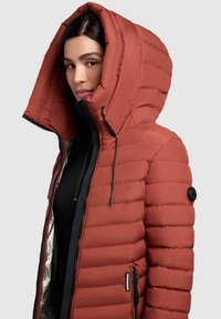 Red puffer jacket with a large hood, ribbed texture, black zipper, and side pockets. Features a logo patch on the left sleeve.