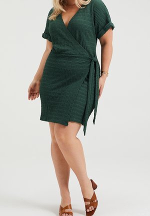 Frau trägt ein strukturiertes, tiefgrünes Wickelkleid mit hochgekrempelten Ärmeln und braunen Sandalen mit Absatz vor neutralem Hintergrund.