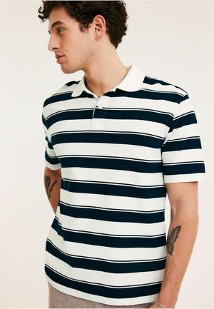 Mann trägt ein kurzärmeliges Polohemd in Marineblau und Weiß gestreift mit Kragen, schaut nach links und hat sichtbare Tattoos auf beiden Armen.