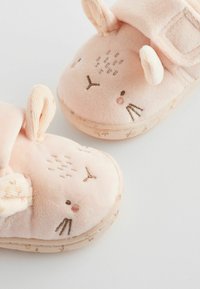 Chaussons pour bébé en peluche rose avec un design de visage de lapin, comportant des détails brodés, des oreilles et un tissu texturé doux. Fermeture à bande Velcro.
