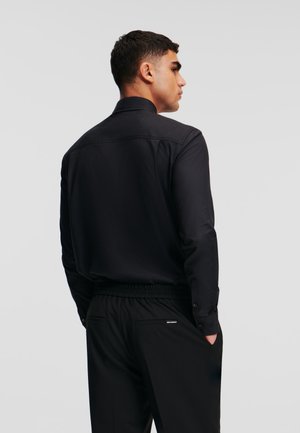 Black long-sleeve shirt with a structured collar, featuring a smooth fabric and button closures, worn with black elastic waistband pants.