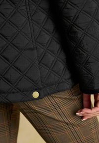 Jachetă neagră matlasată cu un model în romb, având un detaliu cu nasture auriu la tiv; asociată cu pantaloni în carouri în nuanțe de maro și bej.