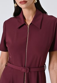 Abito borgogna con maniche corte, chiusura frontale con zip e una cintura coordinata in vita. Tessuto liscio con una leggera lucentezza.