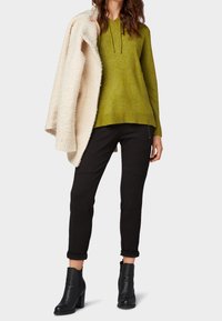 Grüner Hoodie mit Kordelzug, schwarze Hose mit Knieeinnähten und schwarze Stiefeletten mit hohem Absatz. Eingewickelt in einen cremefarbenen, strukturierten Mantel.