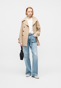 Νεαρή γυναίκα με μπεζ καμπαρντίνα, λευκό πουκάμισο, σκισμένο τζιν ψηλόμεσο, κρατώντας μικρή μαύρη τσάντα, στέκεται μπροστά σε απλό φόντο.