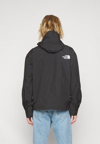 Sort vandtæt jakke med hætte, der har lynlåslukning og logo på den øverste ryg, kombineret med lyseblå jeans.