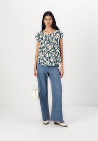Blouse à manches courtes à motif floral dans des tons de vert, crème et bleu, associée à un jean taille haute large bleu clair et des ballerines crème.