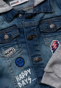 Veste en jean avec boutons, manches de sweat à capuche grises, écussons portant les mots "NOPE", visage triste bleu, éclair coloré et texte "HAPPY DAYS".