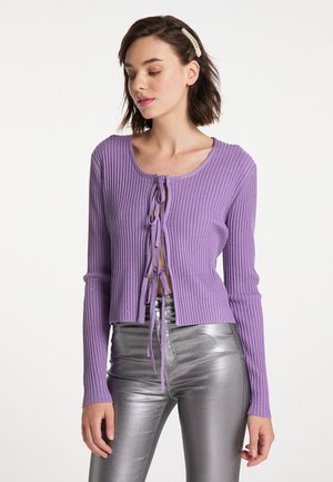Junge Frau trägt eine gerippte violette Strickjacke mit drei Schleifen und metallisch silberne hochtaillierte Hose, schaut nach unten mit sichtbarer Haarspange.