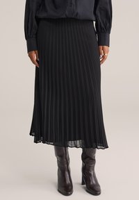 Donna con una gonna midi nera plissettata, una camicia nera con bottoni e stivali in pelle marrone scuro con tacco, in piedi contro uno sfondo neutro.