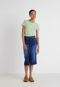 T-shirt vert clair à manches courtes avec texte sur le devant, associé à une jupe en denim mi-longue avec une fente frontale. Chaussures brunes à enfiler.