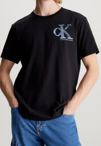 Černé bavlněné tričko s modrou grafickou aplikací "ck" a textem "Calvin Klein Jeans", krátké rukávy a kulatý výstřih.