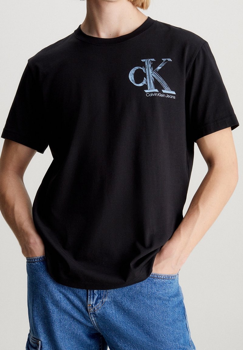 Černé bavlněné tričko s modrou grafickou aplikací "ck" a textem "Calvin Klein Jeans", krátké rukávy a kulatý výstřih.