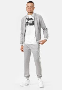 Grijze trainingspak van zacht materiaal, met een ritsjack met strepen, bijpassende broek en een wit T-shirt met een zwart logo. Witte sneakers.