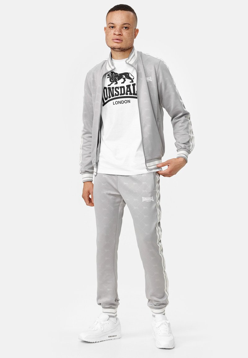 Grijze trainingspak van zacht materiaal, met een ritsjack met strepen, bijpassende broek en een wit T-shirt met een zwart logo. Witte sneakers.