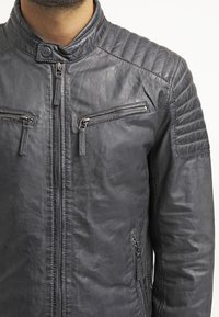 Mørkegrå læderjakke med quiltet skulderdesign, der har to lynlåslommer på brystet og en høj krave. Glat tekstur.