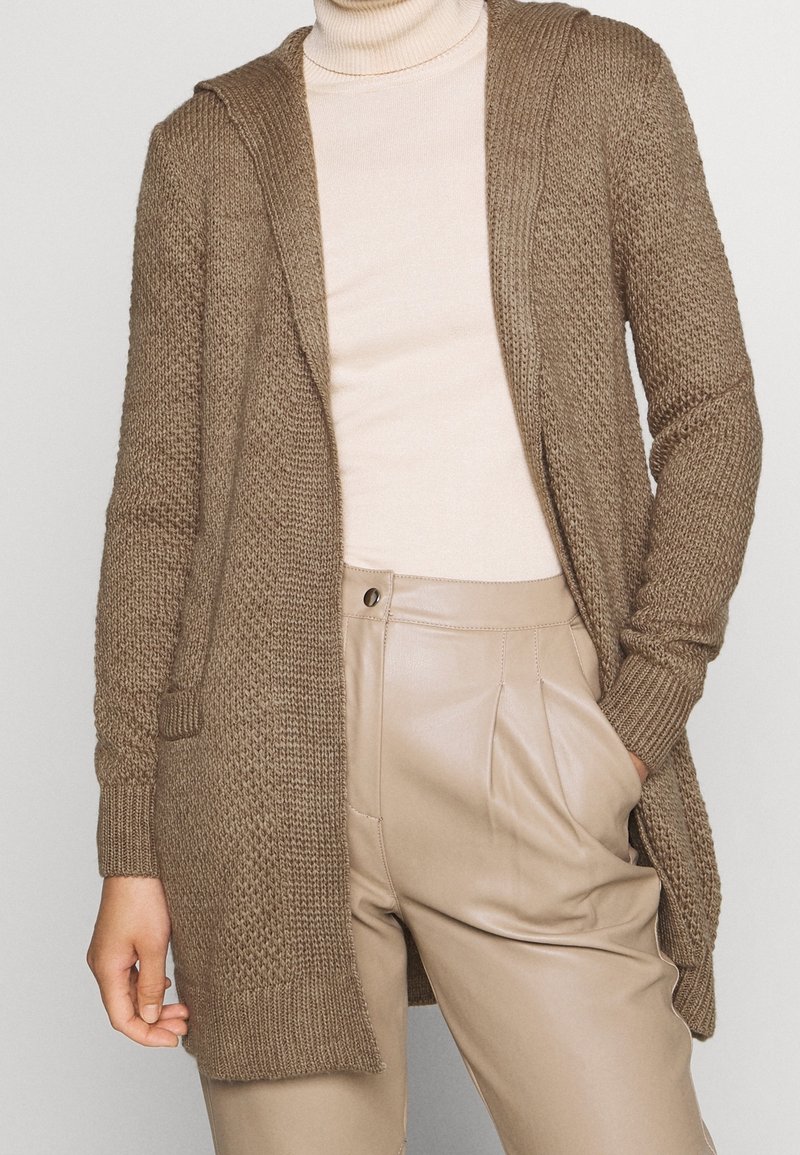 Hnědý pletený cardigan s kapucí, otevřeným předním dílem a žebrovanými manžetami, nošený přes světle béžový rolák, v kombinaci s béžovými elegantními kalhotami.