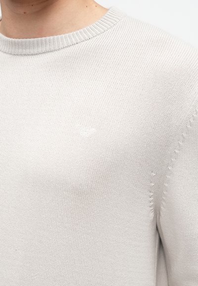 Pull gris clair en tricot avec un col rond côtelé et un discret logo en accent. Présente une texture fine et des détails de couture sur les côtés.