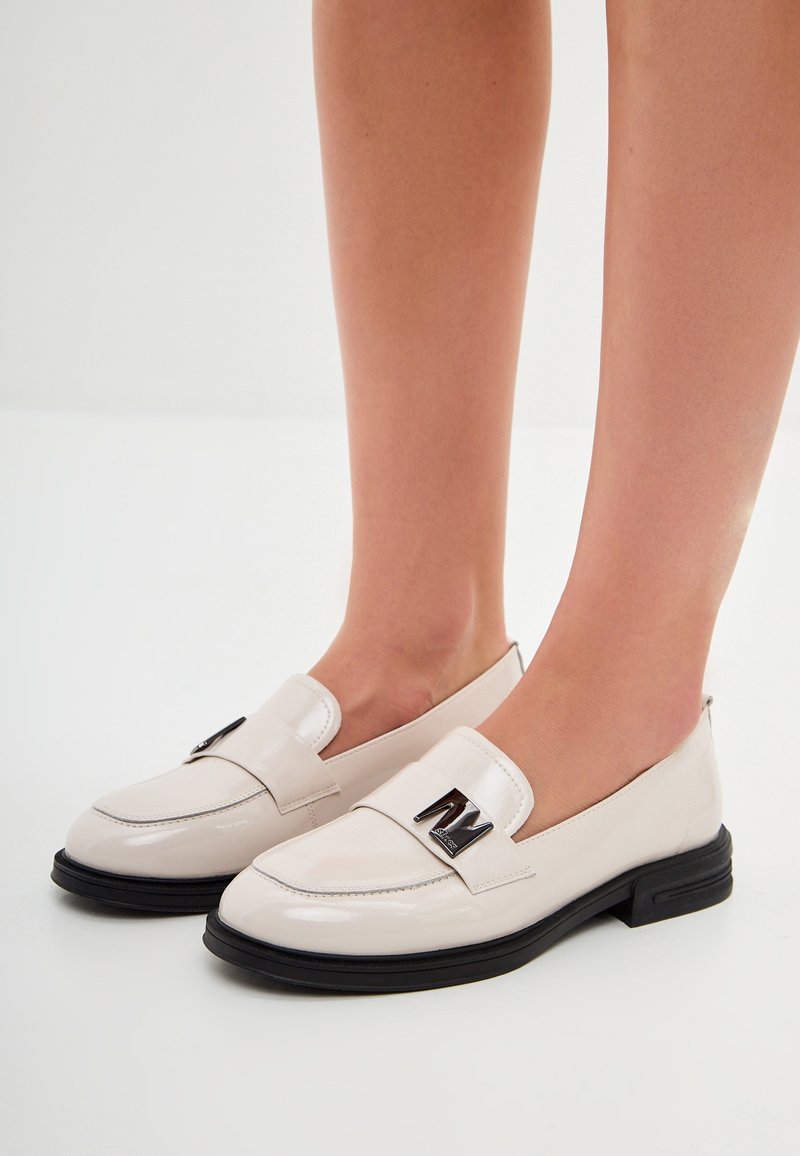 Pieds portant des mocassins en cuir verni crème avec des semelles noires et des boucles décoratives en métal argenté.