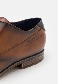 Chaussure en cuir marron avec un bout arrondi et une surface lisse. Présente un talon foncé et un logo "bugatti" en relief. Semelle texturée pour une meilleure adhérence.