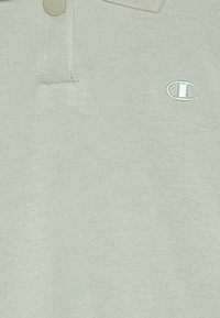 Camisa polo verde-claro feita de tecido de malha, com uma pala de dois botões e um logótipo bordado no peito. Textura suave.