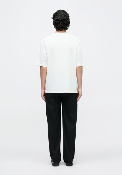 White short-sleeve t-shirt with a round neckline, paired with black trousers and dark shoes, viewed from the back against a plain background.