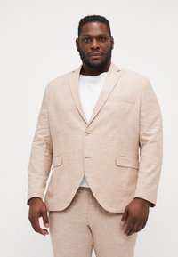 Chaqueta de traje de lino color beige claro con un cierre de un solo botón, solapas de muesca y bolsillos laterales, usada sobre una camiseta blanca.