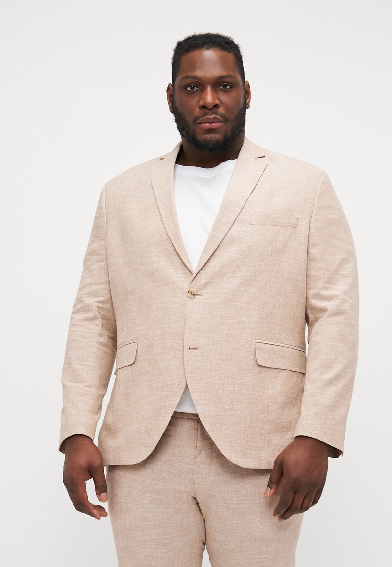 Chaqueta de traje de lino color beige claro con un cierre de un solo botón, solapas de muesca y bolsillos laterales, usada sobre una camiseta blanca.