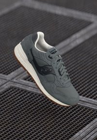 Grå ruskind og mesh sneakers med en tekstureret overdel, hvid mellemsål, brun gummisål og sorte brandingdetaljer på siden.
