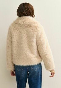 Person med kort brunt hår som bär en ljusbeige fluffig jacka och blå jeans, sedd bakifrån mot en neutral vägg.