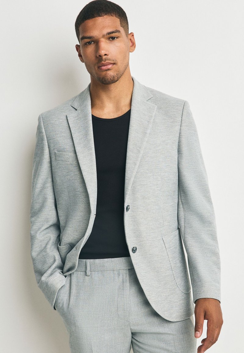 Homme portant une veste de costume gris clair et un pantalon assorti sur une chemise noire, debout avec une main dans la poche devant un fond uni.
