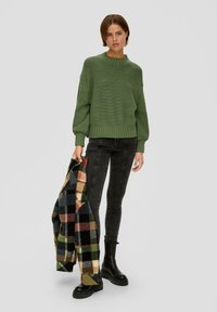 Groene gebreide trui met een geribde halslijn en manchetten, gecombineerd met donkere jeans en zwarte chunky laarzen. Een geruit jasje wordt in de rechterhand vastgehouden.