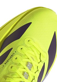 Jasnożółty but do biegania z czarnym wzorem w kształcie łyżwy, teksturowaną cholewką i neonowożółtymi sznurowadłami ściśle związanymi.