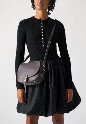 Borsa a spalla in pelle scura con patta, dettagli in metallo dorato e tracolla regolabile, abbinata a un abito nero a coste con gonna ampia.