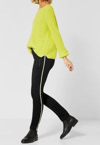 Limenvihreä neulepaita, jossa on väljä istuvuus ja käännetyt hihansuut, yhdistetty mustiin housuihin, joissa on keltainen sivuraidallinen koriste. Mustat nilkkurit.