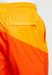 Lehké šortky z hladké látky v oranžové a žluté barvě s diagonálním barevným blokovým vzorem a zadním kapsou na zip.