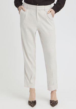 Nainen pukeutuneena luonnonvalkoisiin räätälöityihin housuihin, joissa on käännetyt lahkeet, ja leopardikuvioisiin, kärjestä teräviin korkokenkiin, kädet taskuissa, seisomassa yksivärisen taustan edessä.