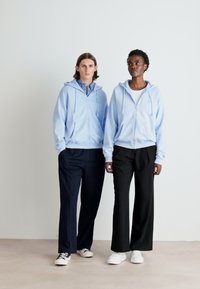 Dos personas llevan sudaderas con cremallera de color azul claro, con bolsillos laterales, combinadas con pantalones oscuros. Las sudaderas tienen cordones y dobladillos acanalados.