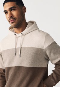 Majica s kapuco z barvnim blokom v bež, sivi in rjavi barvi. Narejena iz mehkega blaga, ima vrvice in kenguru žep.