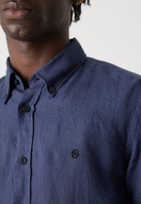 Marineblå knap-up skjorte med en struktureret finish, der har to knapper ved kraven og et lille broderet logo på brystet.