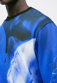 Primer plano de una persona que lleva un sudadera azul con estampados gráficos abstractos en blanco y negro y un cuello de canalé negro.
