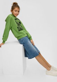 Grüne Kapuzenjacke mit Grafikdruck,blauer Jeansrock und beige Plateausneaker, sitzend auf einem weißen Block vor einem neutralen Hintergrund.